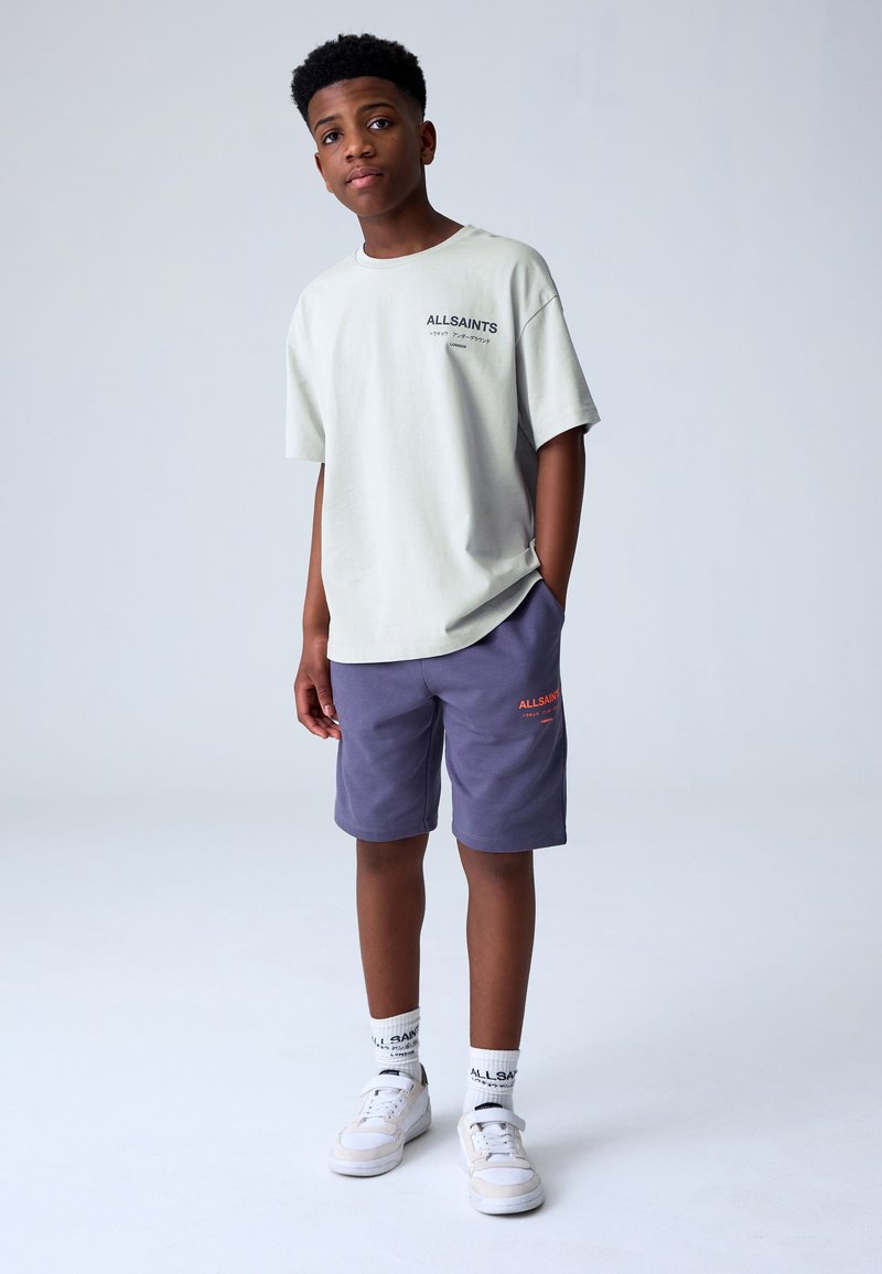 Teenager-Junge steht mit den Händen in den Taschen, trägt ein hellbeiges ALLSAINTS T-Shirt, lila Shorts, weiße ALLSAINTS Socken und weiße Sneakers.