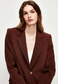 Bruine op maat gemaakte blazer met notch lapels, enkele gouden knoopsluiting en een gestructureerde stof. Bevat een borstzak en gestructureerde schouders.