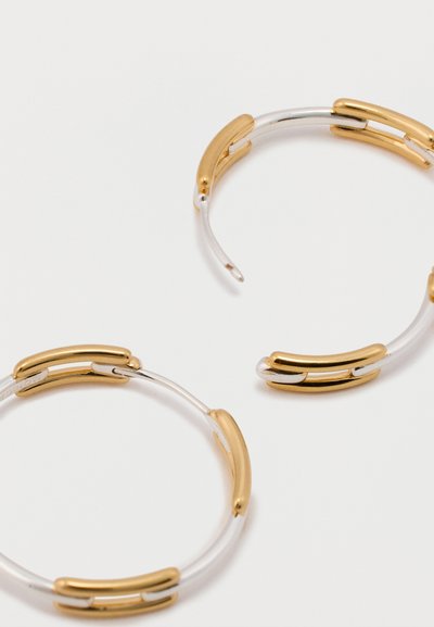 Auksiniai ir sidabriniai auskarai su žiedais, turintys pailgas, sujungtas formas, glotniais paviršiais ir blizgiu apdailu.