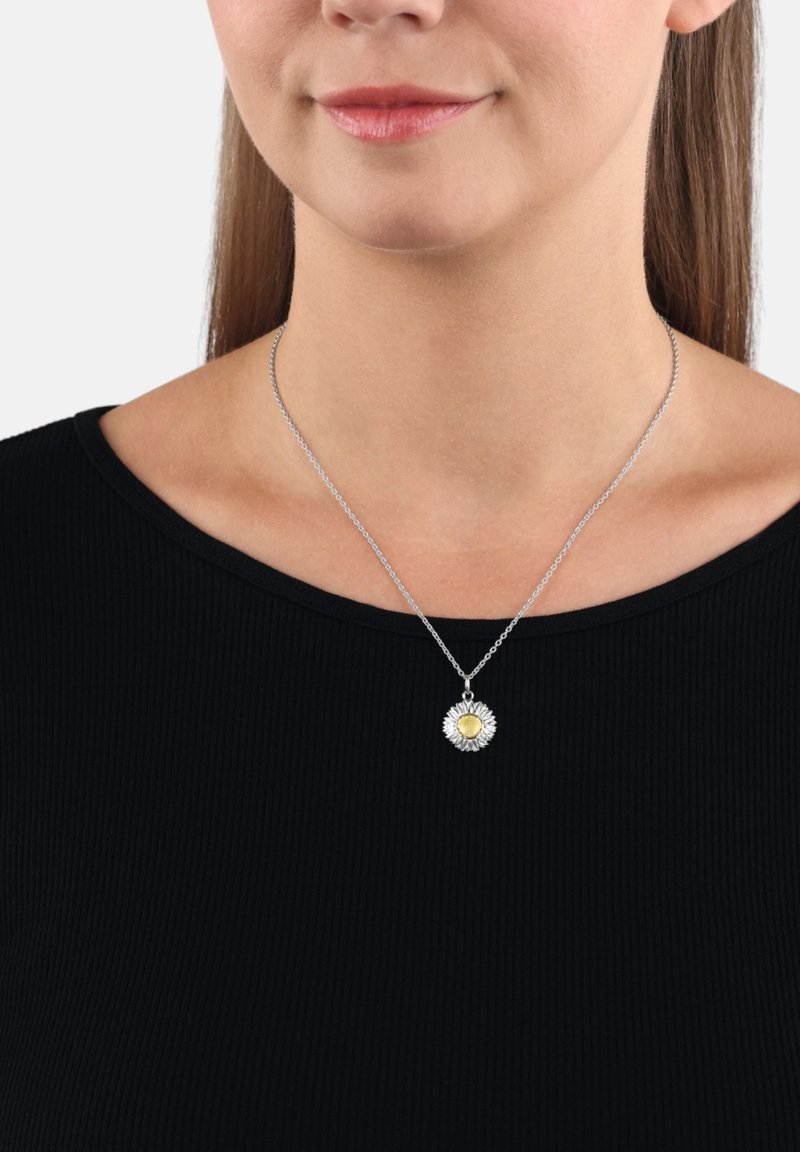 Collana in argento con un pendente rotondo decorato con un motivo floreale, centro giallo e petali in argento texturizzato, indossata sopra una maglietta nera.