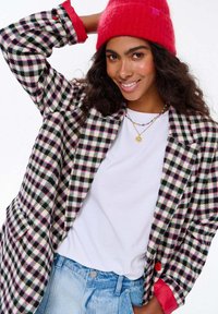 Mladá žena se usmívá, má na sobě červenou čepici, károvanou bundu s růžovými akcenty, bílé tričko, modré džíny a vrstvené barevné náhrdelníky.