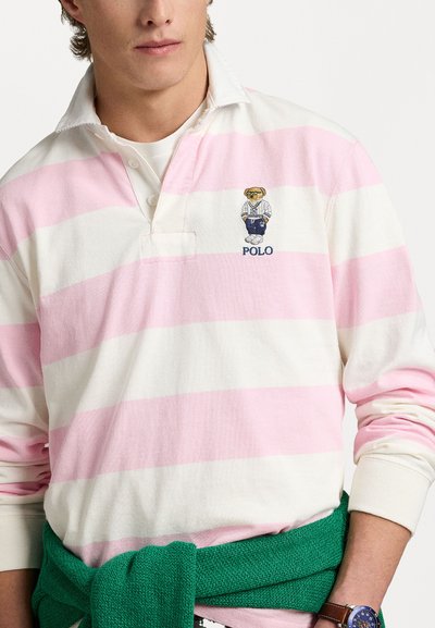 Valge ja roosa triibuline polo särk kraega ning broderitud karu logo. Mudel kannab vöökohtadele seotud roheline sviiter.