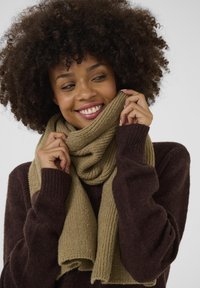 Strikket tørklæde i lys khaki snoet om halsen, vist sammen med en mørkebrun sweater. Strukturen er ribbet; stoffet ser blødt og hyggeligt ud.