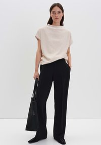 Donna in piedi contro uno sfondo semplice, indossa una maglietta beige a maniche corte, pantaloni neri a gamba larga, scarpe nere e tiene in mano una borsa a tracolla nera.