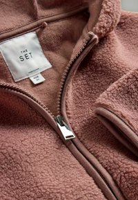 Fleecová bunda v svetloružovej farbe, s manžetou na zips, vysokým golierom a štítkom s označením značky "THE SET." Mäkká textúra a štýlové švy.