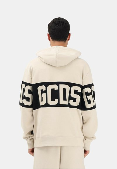 Mies selin, pukeutuneena beigeen huppariin, jossa musta raita ja suuret valkoiset kirjaimet "GCDS" selässä ja hihoissa.