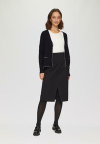 Czarny kardigan z białymi wykończeniami, noszony nad białą bluzką i czarną spódnicą. Czarne rajstopy i loafersy dopełniają look; minimalistyczny design.
