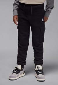 Pantaloni cargo negri de trening, cu buzunare laterale și manșete elastice; prezintă un logo alb pe coapsă. Purtați cu adidași gri și negri.