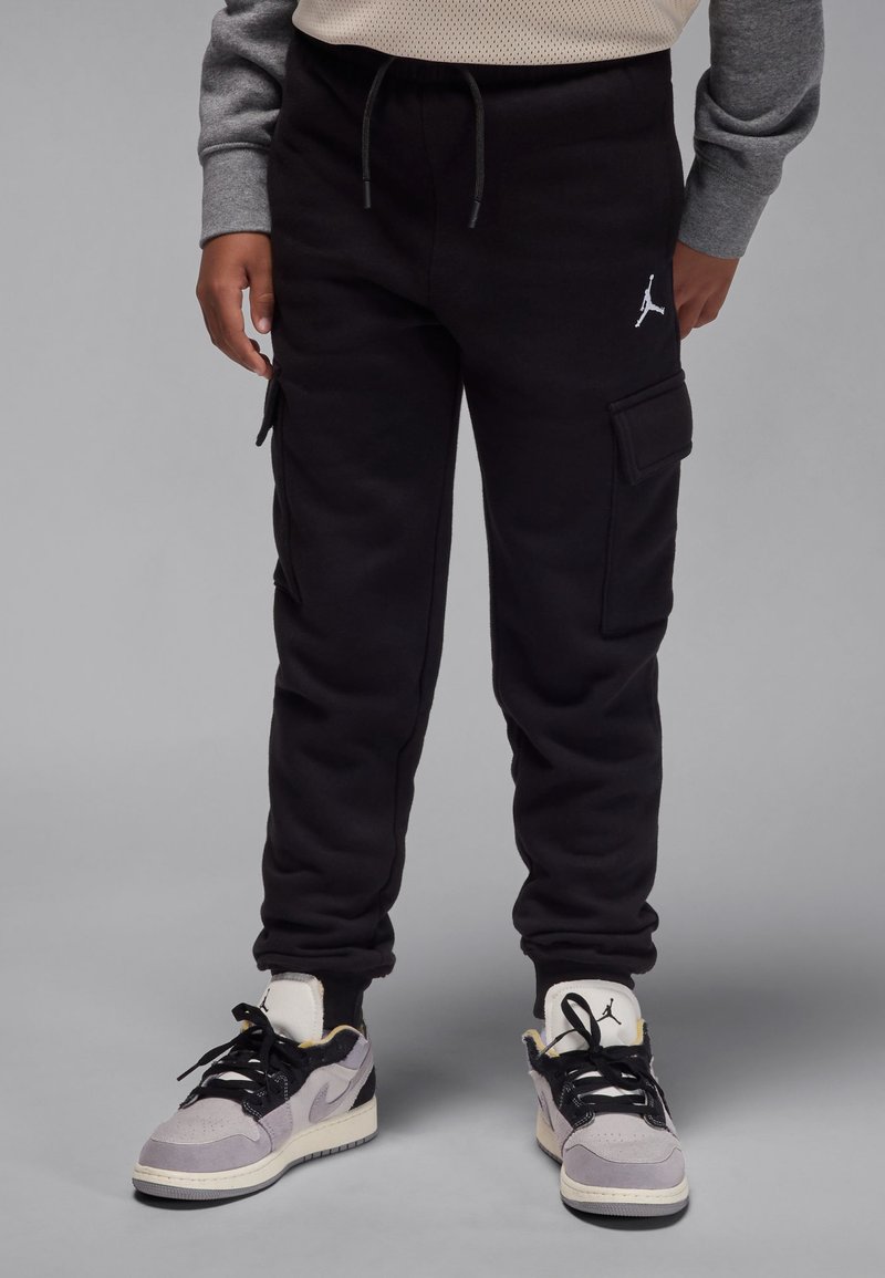 Pantaloni cargo negri de trening, cu buzunare laterale și manșete elastice; prezintă un logo alb pe coapsă. Purtați cu adidași gri și negri.