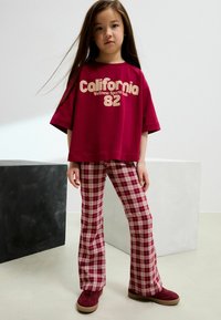 Maglietta in cotone color bordeaux con stampa "California 82"; pantaloni a zampa di elefante a quadretti rossi e bianchi; scarpe in suede marroni; ambiente interno.