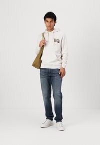 Lys grå hettegenser med frontgrafikk, blå denimjeans og hvite joggesko. Modellen holder en olivengrønn skulderveske.