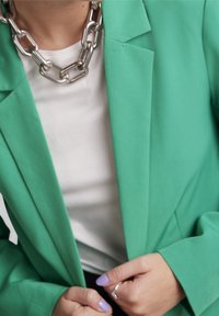 Personne portant une chemise blanche et une veste verte, en train de boutonner la veste, avec un collier en chaîne argentée épaisse et du vernis à ongles violet clair.