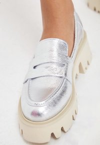Zilveren metallic instappers met een stevige crèmekleurige zool, voorzien van een gladde lederen textuur en een decoratieve band over de wreef.