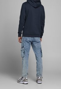 Laivastonsininen huppari, vaaleansiniset cargo-farkut sivutaskuilla ja harmaat lenkkarit valkoisilla yksityiskohdilla. Takaview tasaisella taustalla.