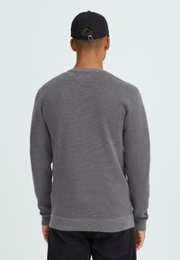 Szürke kötött pulóver texturált mintával, kerek nyakkivágással és bordázott mandzsettákkal és aljjal. Fekete nadrággal viselve, hátulról nézve.