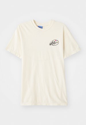 T-shirt de algodão na cor creme, com um decote redondo e mangas curtas. Apresenta um pequeno logotipo em preto e pontos coloridos no lado esquerdo do peito.