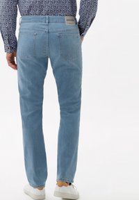 Hombre que lleva jeans de color azul claro, camisa de manga larga con un patrón y zapatillas blancas, mostrado desde atrás contra un fondo blanco.