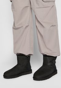 Botas de tobillo de ante negro con forro de piel suave y un detalle de botón, combinadas con pantalones cargo sueltos en gris claro. Suela de goma visible.