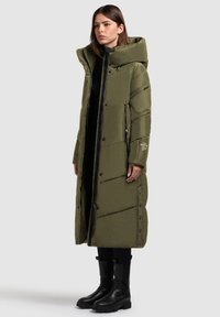 Olivovo zelený dlhý péřový kabát s širohou kapucňou, zipsovým uzáverom, bočnými vreckami a cvokmi. Má prešívaný dizajn a čierne akcenty.