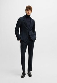 Marinblå kostym med en tvåknappad kavaj och byxor med rak passform, parat med en mörk polotröja. Slät tyg med strukturerad skärning och fickor.