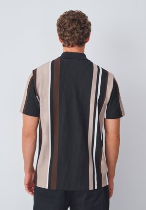 Muž s kudrnatými vlasy, oblečený v krátkém rukávu polo košile s vertikálními černými, hnědými, béžovými a bílými pruhy, viděný zezadu.