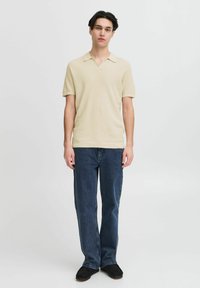 Beige színű, rövid ujjú pólóing V-nyakkivágással, texturált anyagból, sötétkék, egyenes szabású farmernadrággal és fekete cipővel viselve.