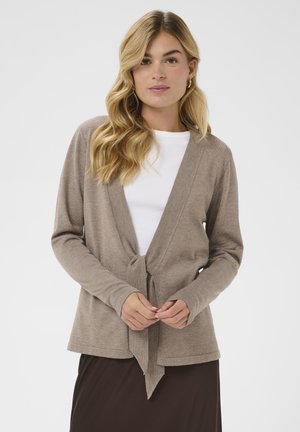 Femme blonde aux cheveux ondulés portant des boucles d'oreilles créoles dorées, un cardigan beige noué devant sur un haut blanc, et une jupe foncée, debout sur fond blanc.
