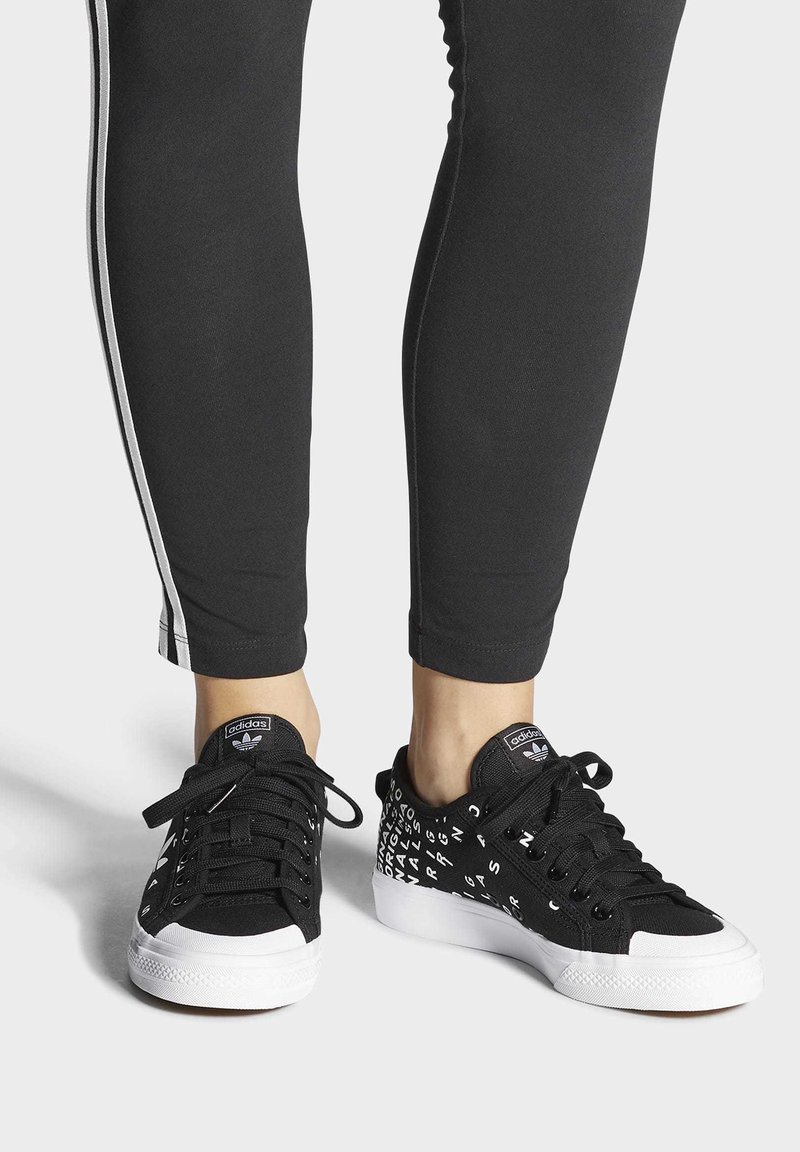 Svarta canvas sneakers med vita gummisulor. Vänster sko har en enhetlig svart design; höger sko visar vita textmönster på en svart bakgrund.