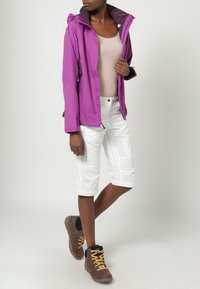 Chaqueta impermeable morada con capucha, pantalones cortos cargo blancos y botas de senderismo marrones. La chaqueta tiene un cierre frontal con cremallera y puños ajustables.