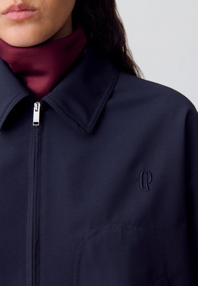 Chaqueta azul marino con cuello, cierre de cremallera y un pequeño logo bordado en el pecho. Textura suave y mate.
