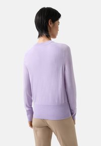 Lila gestrickter Pullover mit geripptem Saum und Bündchen, mit Rundhalsausschnitt und weicher Textur, über beigen Hosen getragen, von hinten betrachtet.