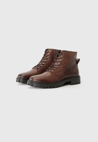 Bottes chelsea à lacets marron avec une semelle robuste, positionnées sur un fond blanc uni. Le cuir semble lisse avec un minimum de détails.