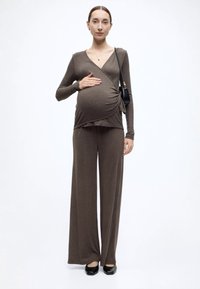 Ένδυμα εγκυμοσύνης από απαλό, ελ αστικό καφέ ύφασμα. Διαθέτει τοπ τύπου wrap με V-λαιμόκοψη και φαρδιά, χαλαρά παντελόνια. Συμπεριλαμβάνονται μαύρα παπούτσια και μικρή τσάντα.