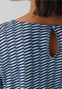 Nahaufnahme des oberen Rückens einer Frau, die ein Oberteil mit geometrischem Muster in Blau und Schwarz trägt, das einen kleinen Ausschnitt und einen einzigen blauen Knopfverschluss hat.