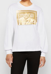 Bílé tričko s dlouhým rukávem, které na přední straně zobrazuje velké zlaté logo Versace Jeans Couture. Tričko má volný střih a žebrovaný kulatý výstřih.