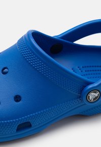Blue clog made of textured plastic, featuring ventilation holes, a back strap, and a logo accent on the side. Smooth, contoured design.