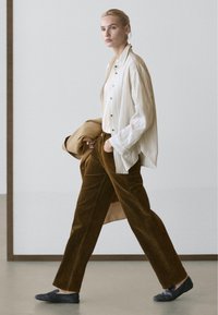 Kvinde går indendørs iført brune kordfløjlsbukser, stribet cremefarvet skjorte, sorte slip-on sko og holder en tanfarvet frakke i venstre hånd.