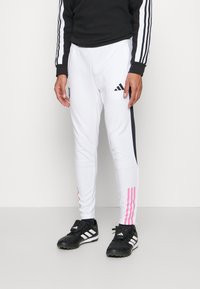 Weisse Sporthosen mit schwarzen Seitenpanelen und pinken Akzenten, gefertigt aus einem glatten Material. Kombiniert mit schwarzen Fußballschuhen mit weißen Streifen.