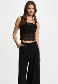 Top corto in pizzo nero con bordi ondulati, dotato di spalline regolabili. Abbinato a pantaloni larghi neri a vita alta.