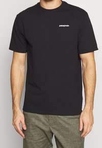Camiseta de algodón negra con mangas cortas, escote redondo y pequeño logo blanco de "Patagonia" en el lado izquierdo del pecho. Ajuste suave y casual.