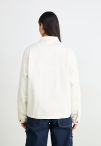 Jachetă din denim alb cu cusături roz, guler și croială lejeră. Partea din spate are o textură netedă și detalii minime.