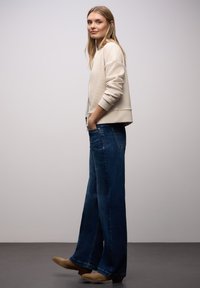 Beige gerippte Jacke, hellgraues Hemd darunter, kombiniert mit weitem blauen Jeans und beige Stiefeletten. Einfaches, lässiges Outfit.