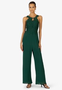 Groene jumpsuit met een bloemenkant lijfje en uitgesneden decolleté. Vloeiende wijde benen, mouwloos ontwerp, aangevuld met zwarte bandjeshakken.