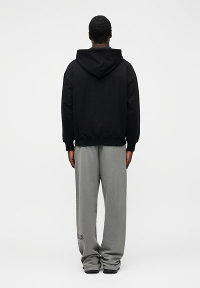 Juodas gobtuvu megztinis su pilkomis sportinėmis kelnėmis, pasižyminčiomis laisvu siluetu, ribotais rankogaliais ir logotipu ant kelnių kojos. Visas paviršius lygus.