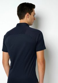 Dunkelblaues Polo-Shirt mit kurzen Ärmeln, aus weichem Stoff, mit Kragen und taillierter Passform sowie dezenten Nahtdetails auf der Rückseite.