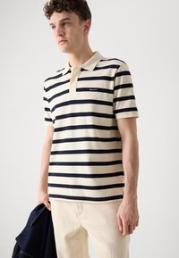 Stripete polo skjorte i krem og marine; mykt bomulls materiale med krage, to-knapps placket, og logo på brystet. Modell holder en jakke.
