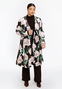 Langer Samt-Cardigan mit floralem Muster in Pink und Grün auf braunem Hintergrund, kombiniert mit dunkelbraunen Hosen und braunen Absätzen.