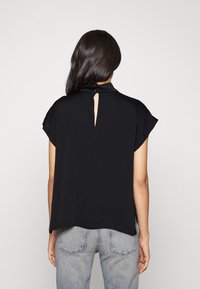 Blusa nera con maniche corte, tessuto texturizzato, leggermente svasata, apertura a chiave sul retro con chiusura a bottone, abbinata a jeans chiari.