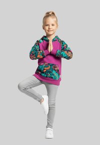 Młoda dziewczyna balansująca na jednej nodze w pozie drzewa, ubrana w fioletową bluzę z kwiatowym wzorem, szare spodnie i białe buty sportowe na szarym tle.