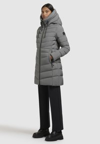Woman wearing a long gray padded hooded coat with hands in pockets, black pants, and black chunky-soled shoes standing sideways.